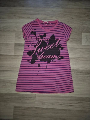 Rosa randig lång topp Sweet Dreams - Kortärmad lång toppi rosa med mörklila ränder och tryck framtill med texten 'Sweet dreams' och svart lövdesign. Mjuk och bekväm, perfekt för mysiga kvällar. Rund halsringning och rak passform.