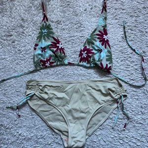 Blommig trekantsbikini med snörning - Snygg bikini med trekantsöverdel i blommigt mönster i grönt, vitt och vinrött. Underdelen är beige med snörningsdetaljer i sidorna. Perfekt för sommaren och strandhäng!