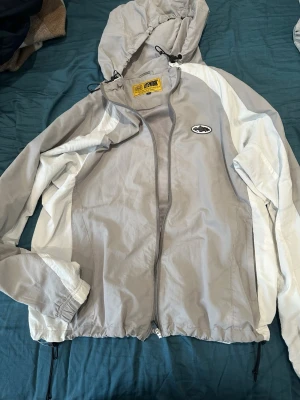 Corteiz gråvit vindjacka med huva - Snygg grå och vit vindjacka från Lemon med dragkedja, justerbar huva och elastiska muddar. Jackan har ett svartvitt logomärke på bröstet och meshfoder på insidan för extra komfort. Perfekt för blåsiga dagar och enkel att matcha med olika outfits.