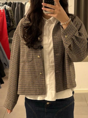 Rutig kappa i bruna toner - Stilren rutig overshirt i bruna och beige toner med klassisk krage och knappar framtill. Jackan har en loose passform och långärmade ärmar, perfekt att slänga över en skjorta för en avslappnad look.
