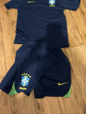 Brasilien Nike fotbollskit marinblå - Snyggt fotbollskit från Nike i marinblått med Brasilien-logga och gula detaljer. Setet består av en t-shirt och shorts med gröna inslag på shortsen. Perfekt för träning eller match och har en sportig passform. Pris diskuteras 