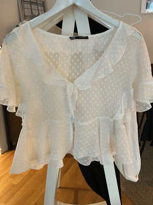 Vit volangblus från Zara - Söt vit blus från Zara med korta ärmar, volangdetaljer och prickigt transparent tyg. Blusen har v-ringning och knappar framtill. Materialet är lätt och ger en romantisk känsla.