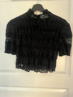 Svart spetsblus - Trendig svart blus i spets med halvtransparenta detaljer. Blusen har volangkant nertill och vid ärmslut samt puffiga korta ärmar. Aldrig anvönd.