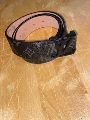 Kappahl bälte  - Snyggt bälte i svart med klassiskt monogrammönster och  i mörk metall. Bältet är gjort i skinn och har en lyxig känsla med beige insida. Perfekt accessoar för att lyfta din outfit.