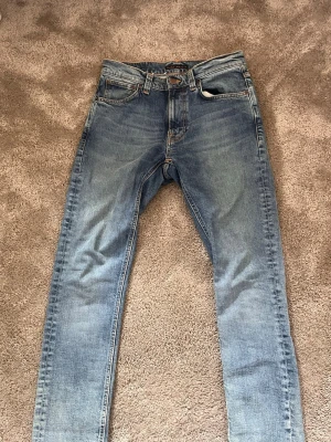Blå jeans från Nudie Jeans Co - Snygga blå jeans från Nudie Jeans Co i klassisk femficksmodell. Jeansen har en tvättad look med ljusa slitningar och raka ben. Tillverkade i slitstarkt denim med orangea sömmar och knappgylf. Perfekta för en avslappnad och trendig stil. Modellen är Lean dean 