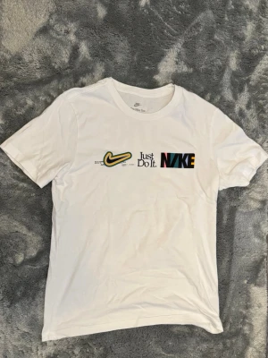Vit Nike t-shirt med tryck M - Säljer en vit t-shirt från Nike med färgglatt tryck fram och bak. Framsidan har Nike-logga, texten 'Just Do It.' och en stor Nike-text i olika färger. Baksidan har stort tryck med text och bilder. Storlek M