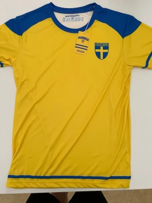 Sverige gul fotbollströja blå detaljer - Gul fotbollströja från Intersport med blå detaljer och Sveriges landslagsemblem på bröstet. Tröjan har rund hals, korta ärmar och blå ränder vid ärmslut och nederkant. Perfekt för att heja på Sverige under matcher.