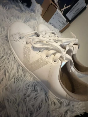 Vita victoria sneakers med vit glitter - Snygga vita sneakers från Victoria med glittrig V-logga på sidan och glitterdetalj vid hälen. Skorna har klassisk låg modell, vita skosnören och ovandel i läder. Perfekta för dig som gillar stilrena men ändå unika sneakers. Aldrig använt