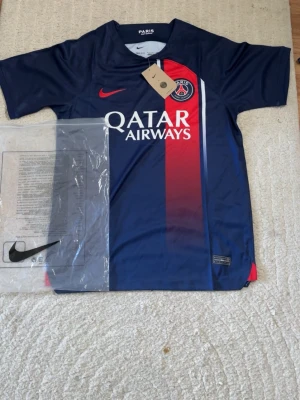 PSG matchtröja Nike blå/röd - Snygg Paris Saint-Germain matchtröja från Nike i blått med röd vertikal rand och vita detaljer. Tröjan har klubbmärke, Qatar Airways-tryck och röd Nike-logga. Tillverkad i lätt och ventilerande Dri-FIT-material, perfekt för fotbollsträning eller match.