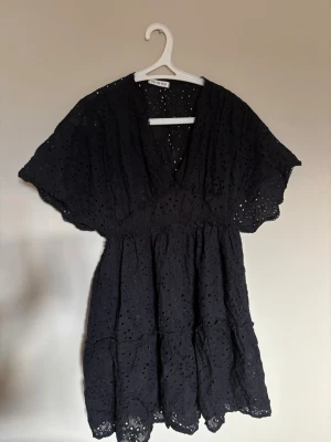 Svart broderad klänning med v-ringning - Supersöt svart klänning från Gina Tricot med v-ringning och broderade detaljer. Klänningen har korta, vida ärmar och resår i midjan för en bekväm passform. Perfekt för dig som gillar romantiska och luftiga plagg.