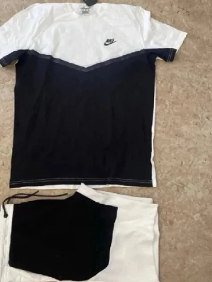 Nike svartvitt set med t-shirt - Snyggt svartvitt set från Nike med en t-shirt i vitt och svart med V-formad färgövergång och Nike-logga på bröstet. Tillhörande shorts i vitt med svart panel och dragsko i midjan. Perfekt för en sportig och avslappnad look.