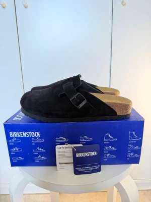 Svarta Birkenstock Boston loafers - Säljer ett par svarta Birkenstock Boston loafers i mocka med justerbart spänne och klassisk korksula. Skorna har en stilren slip-in design och är perfekta för dig som gillar bekväma och trendiga skor. Modellen är tidlös och passar till många olika outfits.