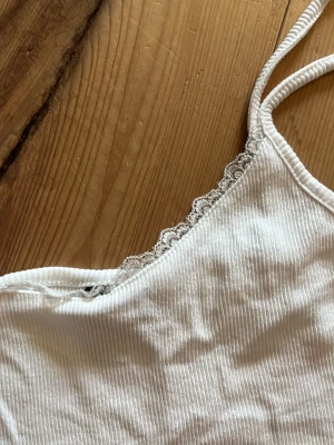 Vit ribbad linnetopp från Vero Moda - Snygg vit ribbad linnetopp från Vero Moda med tunna axelband och spetsdetalj längs urringningen. Perfekt att styla med jeans eller kjol för en fräsch look. Materialet är mjukt och stretchigt för extra komfort.