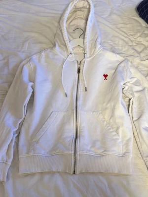 Vit hoodie från Ami Paris - Säljer en clean vit hoodie från Ami Paris med dragkedja, huva med snören och kängurufickor. På bröstet sitter Ami:s ikoniska röda hjärta med A. Tillverkad i mjuk bomull som känns skön mot huden. Perfekt för dig som gillar minimalistisk och stilren design.