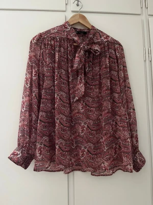 Mönstrad blus med knyt från ZARA - Snygg blus från ZARA med röd och vit paisleymönster, lång ärm och knyt i halsen. Blusen har en lös passform och är tillverkad i ett lätt och luftigt material som passar perfekt till olika stilar.