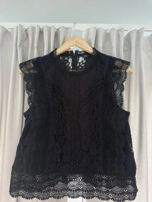 Svart spetsblus från ONLY - Supersnygg svart blus från ONLY med spetsdetaljer och vackert mönstrad kant. Ärmlös modell med rund halsringning och genombruten spets som ger en trendig look. Perfekt att styla med jeans eller kjol för en cool outfit.