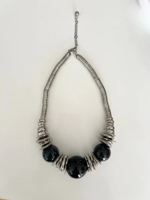 Chunky halsband med svarta pärlor - Stilrent halsband med stora svarta pärlor och många silvriga metallringar. Kedjan är justerbar och består av små, tätt sittande metallbitar. Perfekt för att ge outfiten en cool och modern touch.