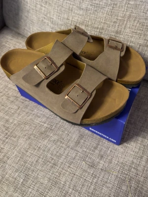 Tofflor  - Snygga beige sandaler  i mjuk mocka med två breda remmar och justerbara metallspännen. Fotbädden är anatomiskt utformad för extra komfort. Perfekta för en avslappnad och trendig stil.