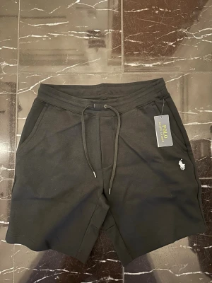 Svarta shorts från Polo Ralph Lauren - Svarta shorts från Polo Ralph Lauren med dragsko i midjan och fickor på sidorna samt en bakficka. Klassisk logga broderad i vitt på ena benet. Tillverkade i mjuk bomull för en bekväm känsla.
