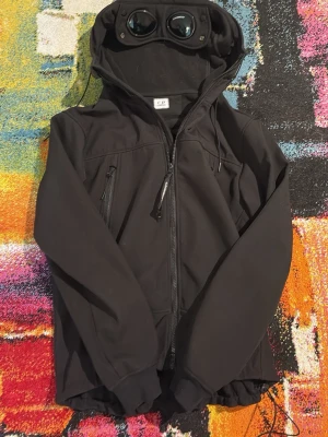  C.P. Company dd shell jacket  - Säljer en svart Goggle Jacket från C.P. Company med ikoniska glasögon i huvan och dragkedja framtill. Jackan har två snedställda bröstfickor med dragkedja och elastiska muddar. Perfekt för dig som vill sticka ut med en unik och funktionell jacka.