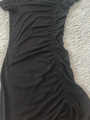 Svart kortklänning med snörning - Snygg svart kortklänning med snörning längs sidan som ger en cool rynkad effekt. Klänningen har korta ärmar och är tillverkad i ett mjukt, stretchigt material som sitter bekvämt. Perfekt för dig som gillar trendiga detaljer och enkel stil.