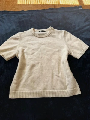 Beige stickad topp med kort ärm - En enkel och stilren beige stickad topp med rund hals och korta ärmar. Ribbstickade kanter vid ärmslut, halsringning och nederkant ger en clean look. Perfekt att matcha med jeans eller kjol för en chill vibe. Storlek M från Gina tricot
