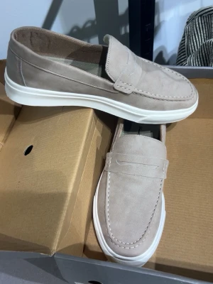 Beige loafers med vit sula - Snygga beige loafers med klassisk penny-detalj och vit, räfflad sula. Skorna har en stilren design med mjukt material och diskreta sömmar runt tån. Perfekta för dig som gillar en clean och modern look. perfekt för din student/bal 