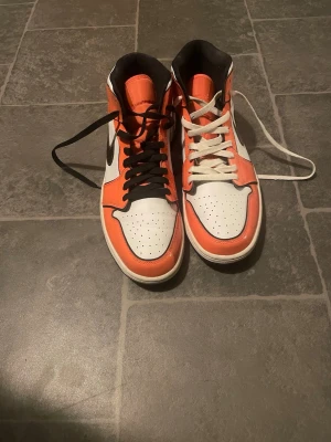 Air Jordan 1 Orange/White/Black Highs - Säljer ett par Air Jordan 1 med högt skaft i orange, vitt och svart. Klassisk Nike Swoosh på sidan, perforerad tåbox och kontrasterande svarta och vita skosnören. Skorna har läderöverdel och ikonisk Air Jordan-logga vid ankeln.