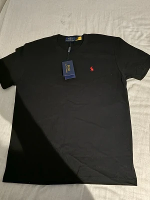 Svart t-shirt från Polo Ralph Lauren - Svart t-shirt från Polo Ralph Lauren med klassisk rund hals och röd broderad logga på bröstet. Enkel och stilren design som passar till det mesta. Tillverkad i mjuk bomull för en skön känsla.
