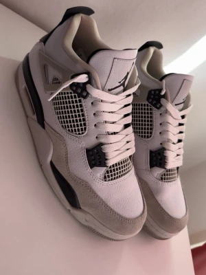 Air Jordan 4 Retro Vit/Svart/Grå - Säljer ett par Air Jordan 4 Retro sneakers i vitt, svart och grått. Skorna har meshpaneler, mockadetaljer och klassisk Jumpman-logga på hälen. Tjock sula och ikoniska plastdetaljer vid snörningen ger en sportig vibe. Perfekta för dig som gillar streetwear och snygg design.
