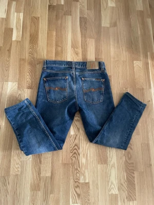Blå jeans från Nudie Jeans - Säljer ett par klassiska blå jeans från Nudie Jeans med raka ben och snygga kontrastsömmar. Jeansen har två bakfickor med Nudie Jeans signaturbrodyr och en patch i läder bak i midjan. Perfekta för en avslappnad och stilren look. Storlek: w27 l30 men sitter lite mindre i längden!