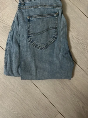 Ljusblå Lee jeans med bakficka - Säljer ett par klassiska ljusblå jeans från Lee med en snygg bakficka och detaljerade sömmar. Jeansen har normal midja och raka ben, tillverkade i slitstarkt denimtyg. Perfekta för dig som gillar tidlös och avslappnad stil.