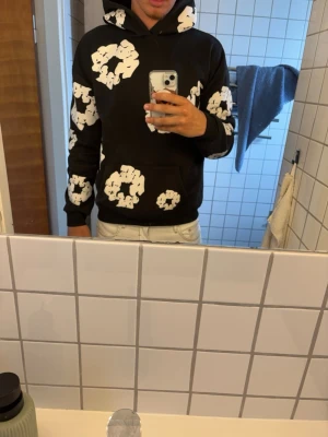 Svart hoodie med denim tears  - Cool svart hoodie med stora vita blommönster över hela tröjan. Klassisk passform med huva och känguruficka framtill. Perfekt för dig som vill sticka ut med en unik och trendig stil.