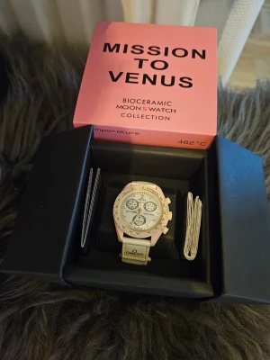 Omega x Swatch Mission to Venus klocka - OBS. boxen är trasig! Säljer en Omega x Swatch Mission to Venus från Bioceramic MoonSwatch Collection. Klockan har en ljusrosa boett, vit urtavla med flera subdials och tachymeter-skala. Armbandet är ljust med Omega-logga. En riktigt snygg och unik accessoar för dig som gillar statement pieces.