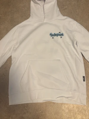 Vit hoodie från Hoodrich med tryck - Säljer en vit hoodie från Hoodrich med blått och svart graffiti-inspirerat tryck på bröstet och ryggen. Hoodien har en stor magficka och klassisk huva. Perfekt för dig som gillar streetwear och vill sticka ut med coola detaljer.
