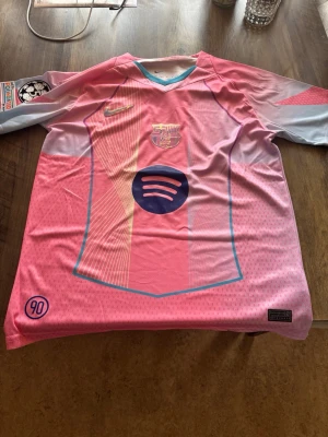 Rosa FC Barcelona matchtröja Nike - Säljer en rosa FC Barcelona matchtröja från Nike med klubbmärke och Spotify-logga på bröstet. Tröjan har ljusblå och gula detaljer, korta ärmar och är tillverkad i ett lätt, ventilerande funktionsmaterial. Perfekt för fotbollsträning eller att bära till vardags.