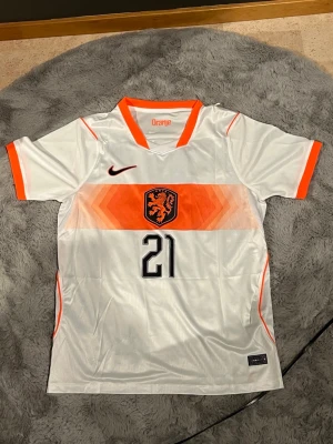 Vm De Jong Number 21 fotbollströja  - Snygg vit Holland fotbollströja från Nike med orange detaljer, nummer 21 och De Jong på ryggen. Tröjan har V-ringning, korta ärmar och ett stort lejonmärke på bröstet. Perfekt för dig som älskar fotboll och vill stötta Nederländerna.