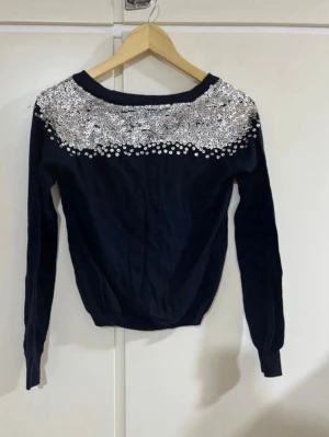 Svart kofta med paljetter från Gina Tricot - Snygg svart kofta från Gina tricot med långa ärmar och silverfärgade paljetter över axlar och bröst. Koftan har v-ringning och knäppning framtill. Perfekt för dig som vill sticka ut lite extra med glittriga detaljer.