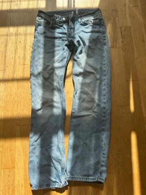 Blå raka jeans med slitningar - Säljer ett par blå jeans med rak passform och lätt slitna detaljer. Jeansen har klassisk femficksdesign och knappgylf. Perfekta för en avslappnad och trendig look.