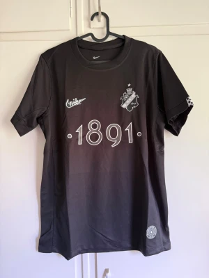 Svart AIK x Nike matchtröja - Snygg svart AIK matchtröja från Nike med trycket '1891' på bröstet och klubbmärke. Tröjan har rund hals, korta ärmar och texten 'SMOKINGHJÄRTA' i nacken. Tillverkad i lätt och ventilerande material, perfekt för träning eller match.