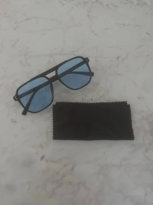 Blåa solglasögon  - Snygga solglasögon med svarta pilotbågar och blåtonade glas. Klassisk aviator-modell som ger en cool vibe. Medföljer en svart putsduk. Perfekta för soliga dagar och trendiga outfits. Helt nya alltså oanvända!