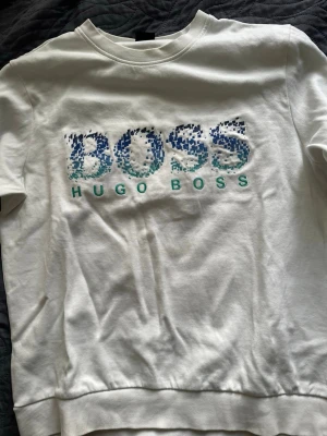 Vit sweatshirt från Hugo Boss - Snygg vit sweatshirt från Hugo Boss med stort BOSS-tryck i blått och grönt framtill. Tröjan har rund halsringning, ribbade muddar och är tillverkad i mjukt bomullsmaterial. Perfekt för en avslappnad och trendig look. Pris kan diskuteras.