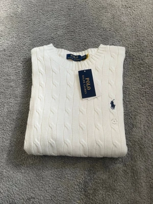 Vit kabelstickad tröja Polo Ralph Lauren - Vit kabelstickad tröja från Polo Ralph Lauren med rund halsringning och klassisk blå logga broderad på bröstet. Tröjan har ribbade muddar och är gjord i mjukt material som passar perfekt till lager-på-lager-stil. Har provat tröjan en gång utan att ha använd den under vardagen. Finns ett litet ursprungligt fel