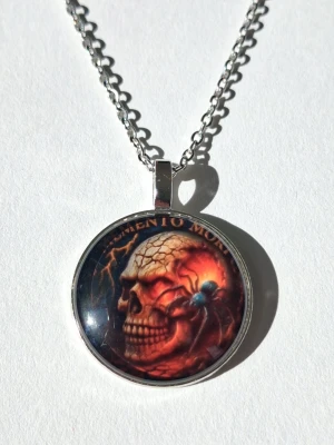 Halsband med dödskalle och spindel - Snyggt halsband med runt hängsmycke I 2D som har ett motiv av en dödskalle och en blåsvart spindel. Texten 'Memento Mori' syns upptill. Kedjan är i silverfärgad metall och hänget har en glansig yta. Perfekt för dig som gillar alternativ stil eller vill sticka ut. Materialet rostfritt stål. 