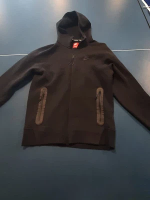 Svart Nike hoodie med dragkedja - Svart hoodie från Nike med hel dragkedja framtill och två fickor med dragkedja. Klassisk huva och diskret Nike-logga på bröstet. Perfekt för en sportig och avslappnad stil. Materialet är mjukt och bekvämt, passar till många tillfällen.