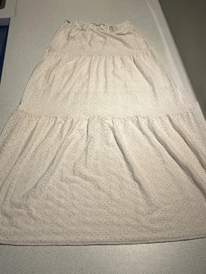 Vit långkjol med broderade detaljer - Söt vit långkjol med broderade hålmönster och volang. Kjolen har resår i midjan och är perfekt för varma dagar. Lätt och luftig modell som ger en romantisk känsla.