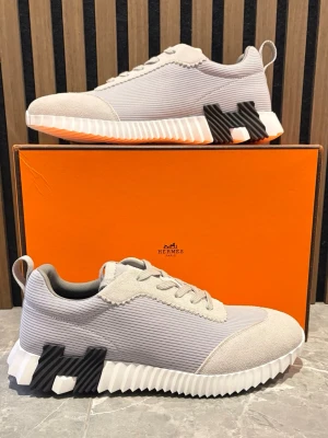  grå sneakers med H-detalj - Snygga sneakers i ljusgrå mesh och mocka med ikonisk H-detalj på sulan. Skorna har vita, räfflade sulor och matchande grå skosnören. Perfekta för dig som vill ha en stilren och exklusiv look med en modern twist.