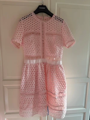 Rosa spetsklänning från By Malina - Söt rosa kortklänning från By Malina med rund halsringning och korta ärmar. Klänningen har ett genombrutet spetsmönster, markerad midja och dragkedja bak. Perfekt för sommarens alla tillfällen.
