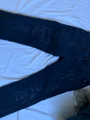Svarta Replay jeans med slitningar - Snygga svarta jeans från Replay i modellen Anbass. Jeansen har coola slitningar och patchade detaljer på benen för en trendig look. Klassisk femficksmodell med knappstängning och bälteshällor. Perfekta för dig som gillar streetstyle.