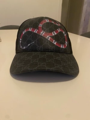 Svart keps med ormtryck - Snygg svart keps  med klassiskt GG-mönster och en stor röd och vit orm på framsidan. Kepsen har mesh på sidorna och justerbar rem bak. Perfekt för dig som vill sticka ut med en exklusiv accessoar.